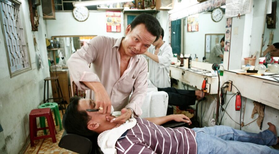 A barber shaving a reclining client with a straight razor in a barbershop.